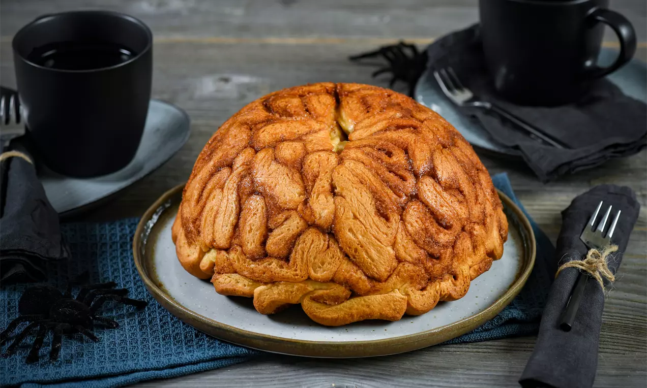 Gehirnförmiger Kuchen mit braun-goldener Oberfläche auf einem Teller, arrangiert mit schwarzen Tassen, Servietten und dekorativen Spinnen