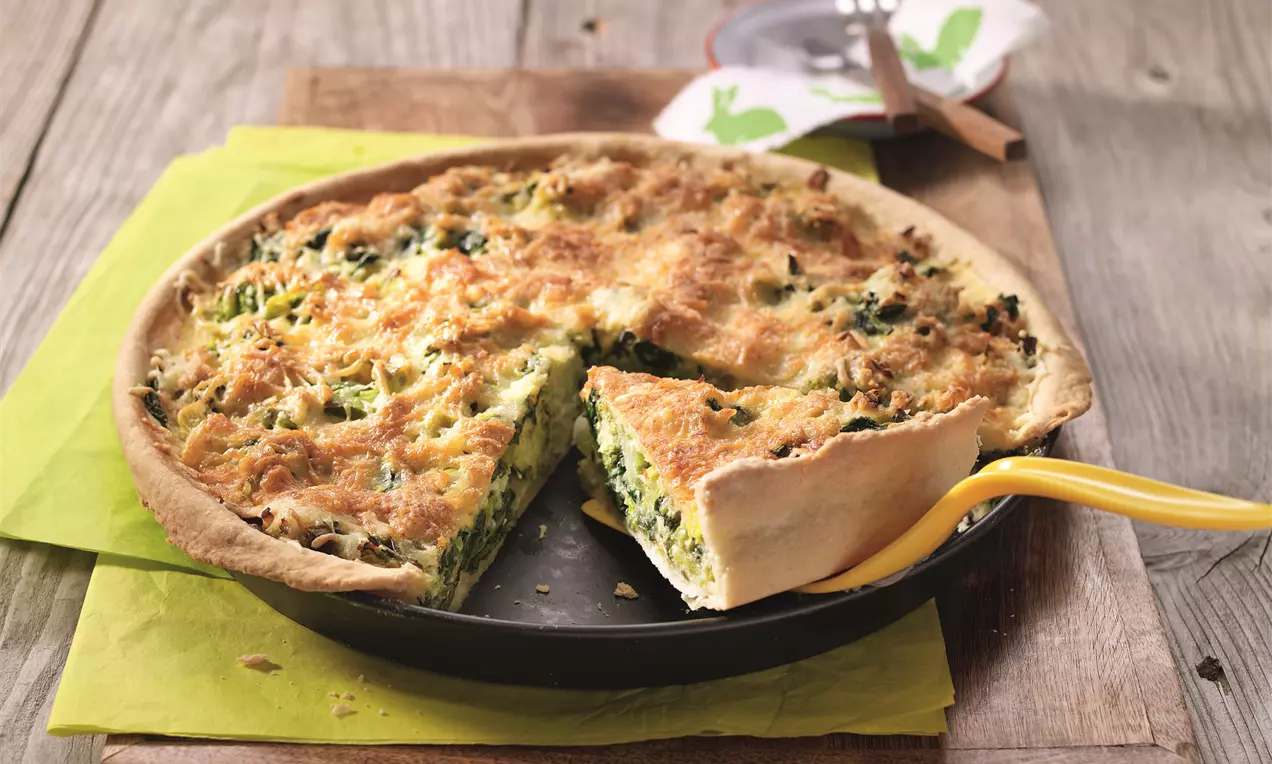 Goldbraune Wähe mit grüner Füllung aus Gemüse und Käse in schwarzer Backform, angeschnitten, auf einem Holztisch mit gelber Serviette drapiert