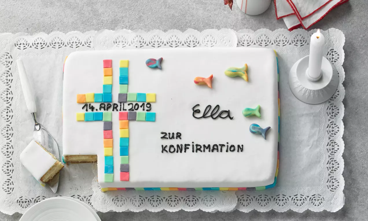 Rechteckige Torte mit weißem Fondant, buntem Mosaikkreuz, Fischen aus Zucker, Schriftzug und einem Anschnitt auf Spitzendeckchen drapiert