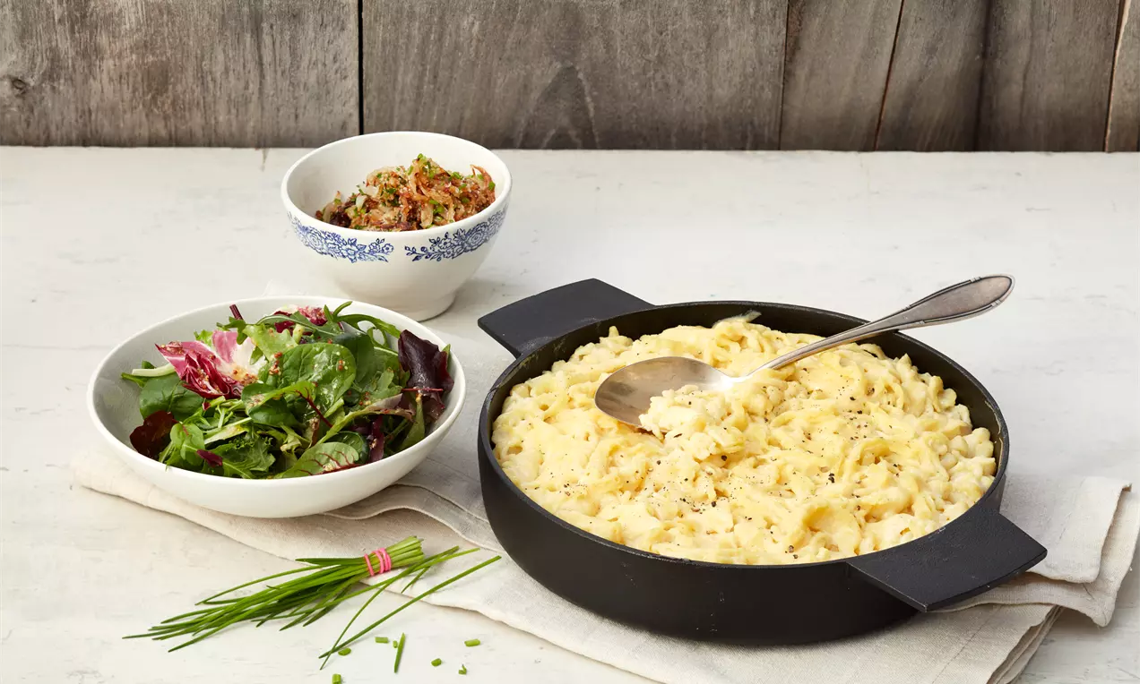 Goldgelbe Käsespätzle in schwarzer Pfanne, cremig und mit Kräutern bestreut, daneben Salatschale und kleine Schüssel auf hellem Tisch