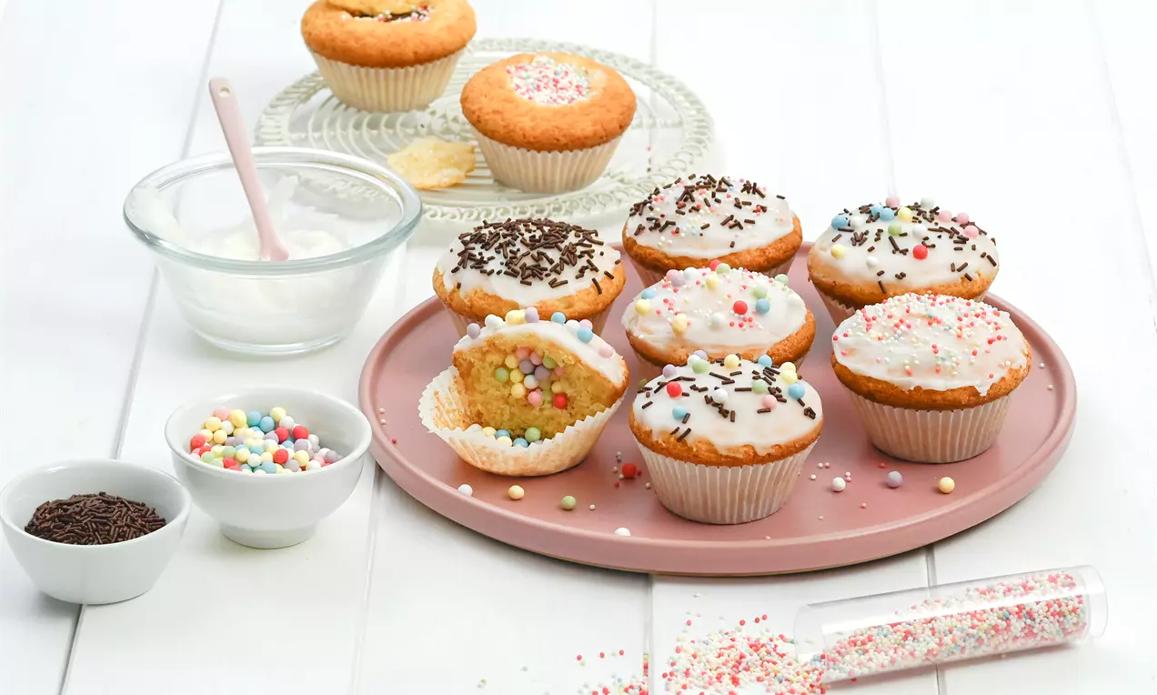 Bunte Piñata-Muffins mit Zuckerguss und Streuseln auf rosa Teller, ein angeschnittener Muffin zeigt farbige Perlen innen, umgeben von Zutaten