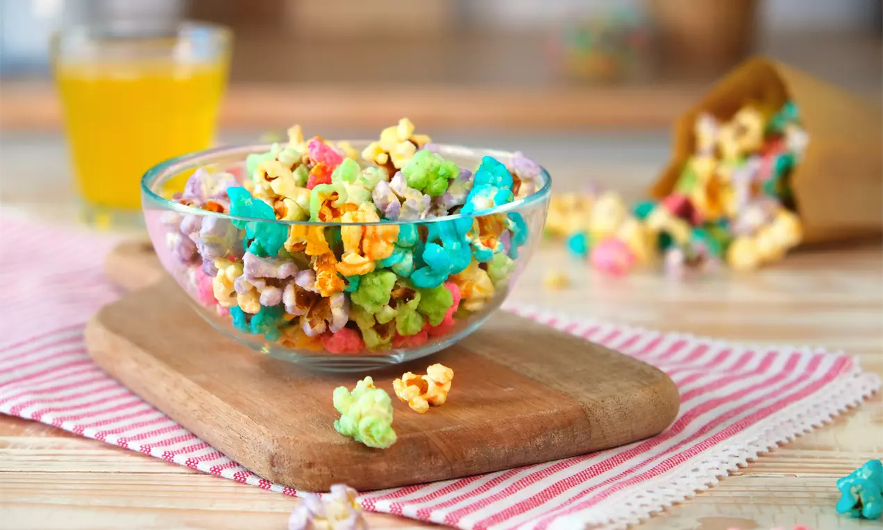 Bunt glasiertes Popcorn in kräftigen Farben wie Blau, Grün und Gelb in einer Glasschale, arrangiert auf einem Holzbrett mit Tuch drapiert