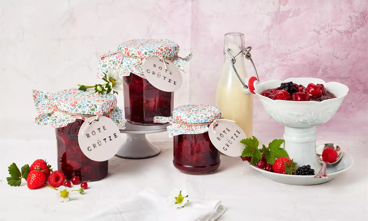Eingekochte rote Grütze in gläsernen Marmeladengläsern mit bunter Haube, arrangiert mit frischen roten Beeren und weißer Milchflasche