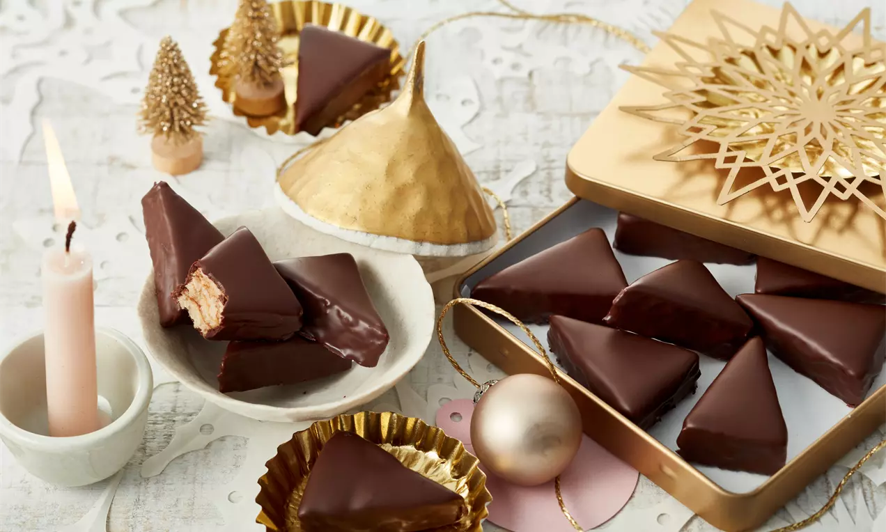 Schokoladenüberzogene Baumkuchenspitzen in dreieckiger Form, teils angeschnitten, auf hellem Teller mit goldener Verpackung und Weihnachtsdekor