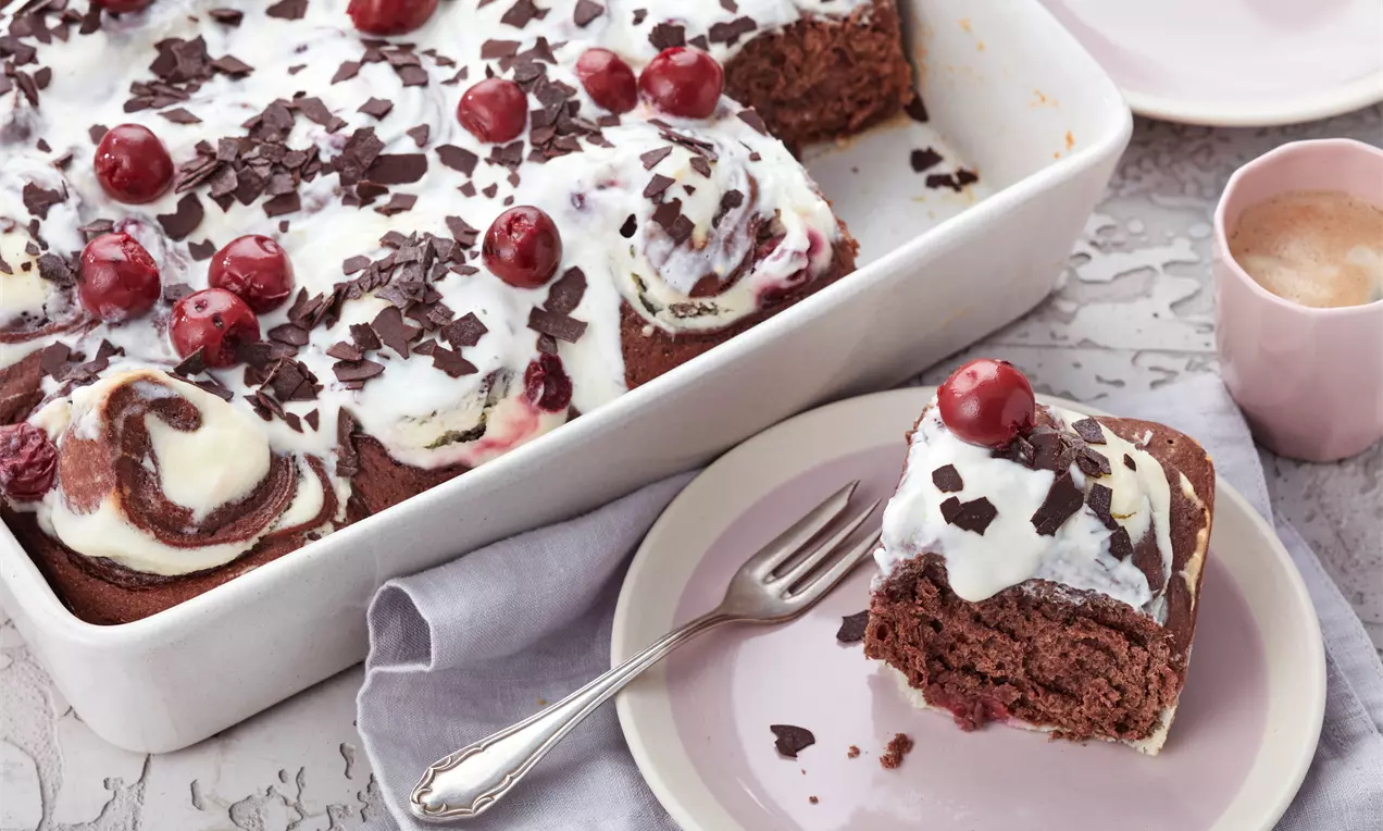 Schwarzweiß marmorierte Schokorollen mit weißer Creme, roten Kirschen und dunklen Schokoraspeln in weißer Form, ein Stück auf rosa Teller