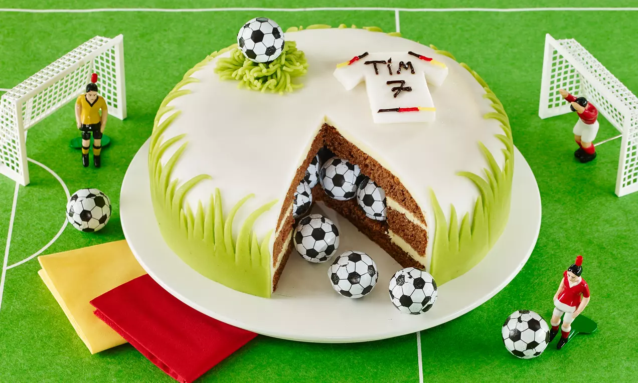 Runde Torte mit weißem Fondant, grünem Grasmuster und Fußballdekor, angeschnitten mit gefüllten Schokokuchen und kleinen Fondant-Bällen