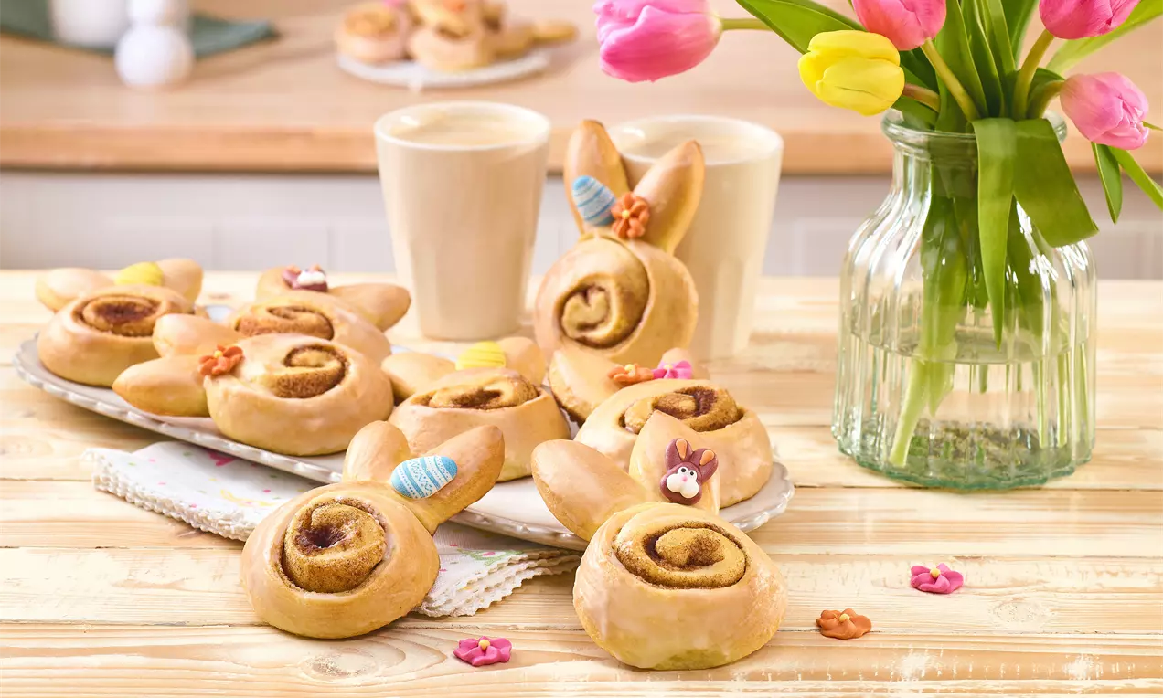 Zimtschnecken in Osterhasenform mit Teigohren und kleinen Zuckerdekos, auf einem Holztisch angerichtet; dazu zwei Tassen Kaffee und eine Vase mit bunten Tulpen im Hintergrund.