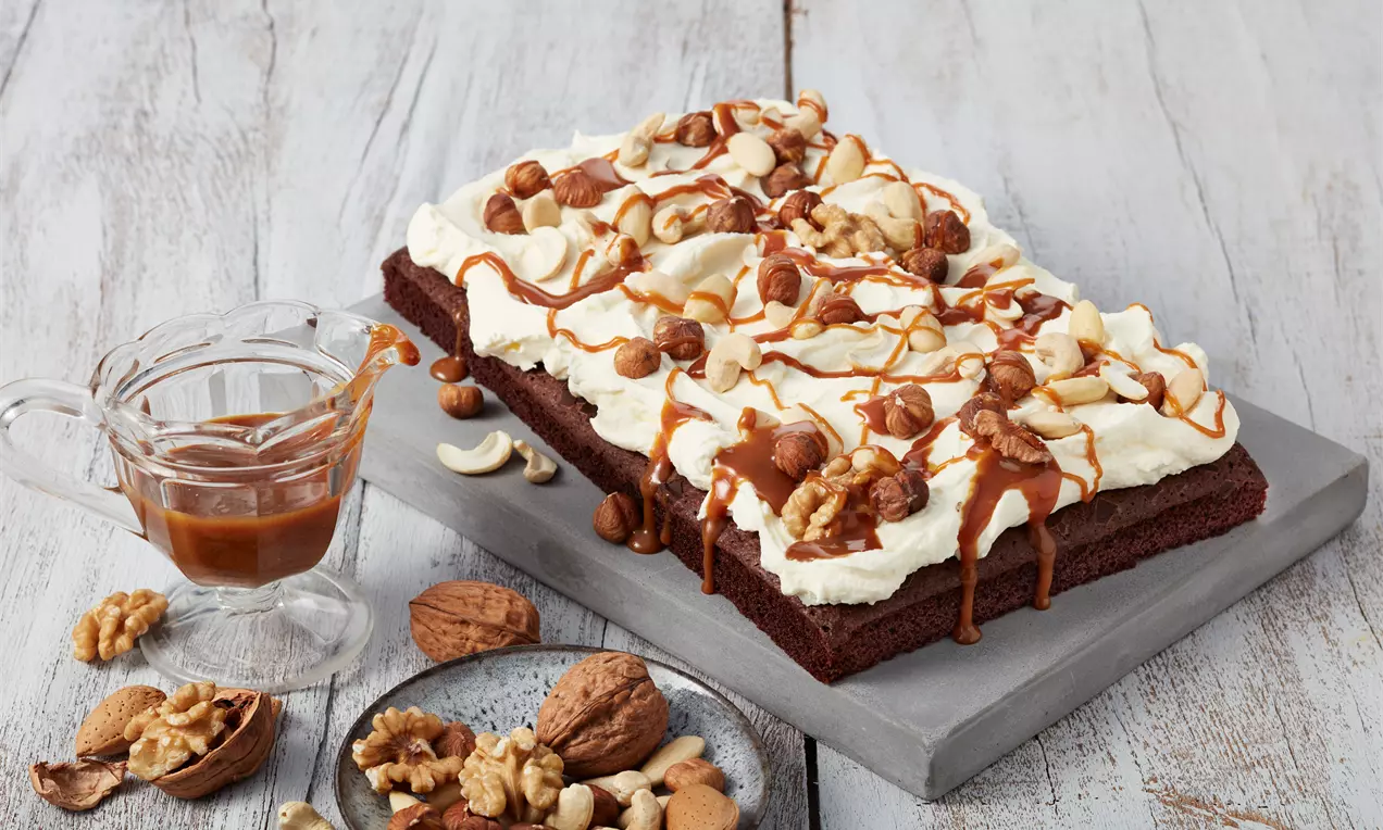 Rechteckige Brownies mit cremeweißer Frischkäse-Schicht, garniert mit Nüssen und Karamellsoße auf grauem Untergrund angerichtet