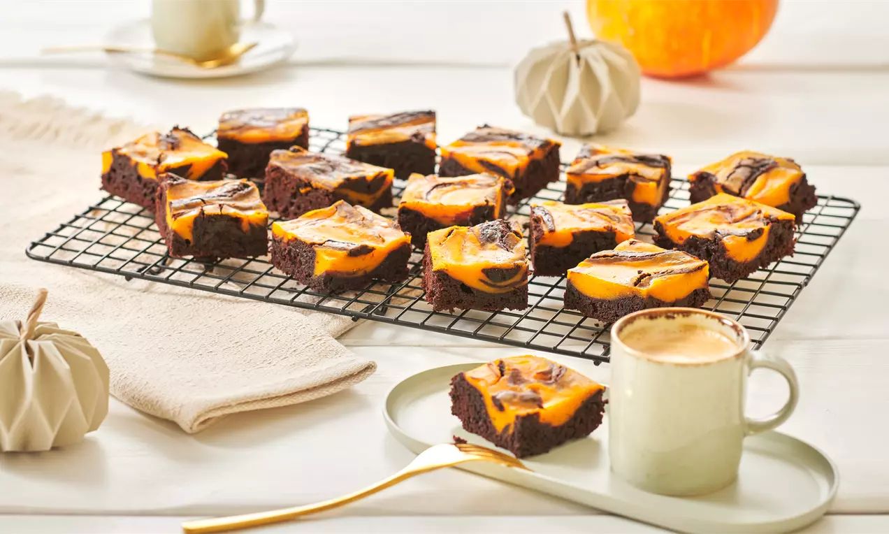 Saftige Kürbis-Brownies mit orange-gelb marmorierter Käsekuchenschicht auf einem schwarzen Gitterrost, arrangiert mit Kürbisdeko und Kaffee