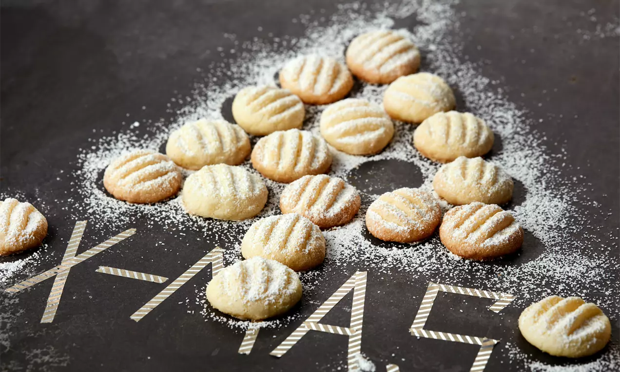 Kleine, goldgelbe Schneeflöckchen mit feinem Puderzucker bestäubt, kreisartig angeordnet auf dunkler Oberfläche mit X-MAS-Schrift