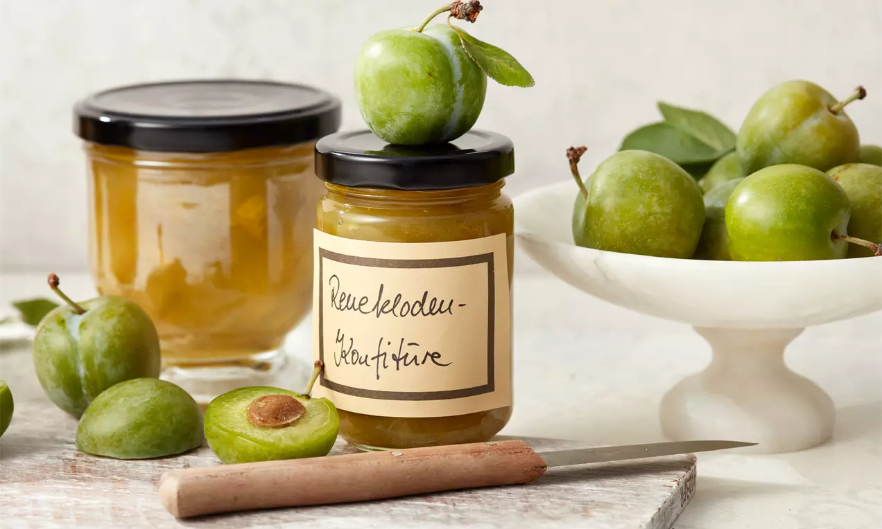 Renekloden-Konfitüre im Glas mit handgeschriebenem Etikett, umgeben von grünen Früchten und einem angeschnittenen Renekloden auf Holzbrett
