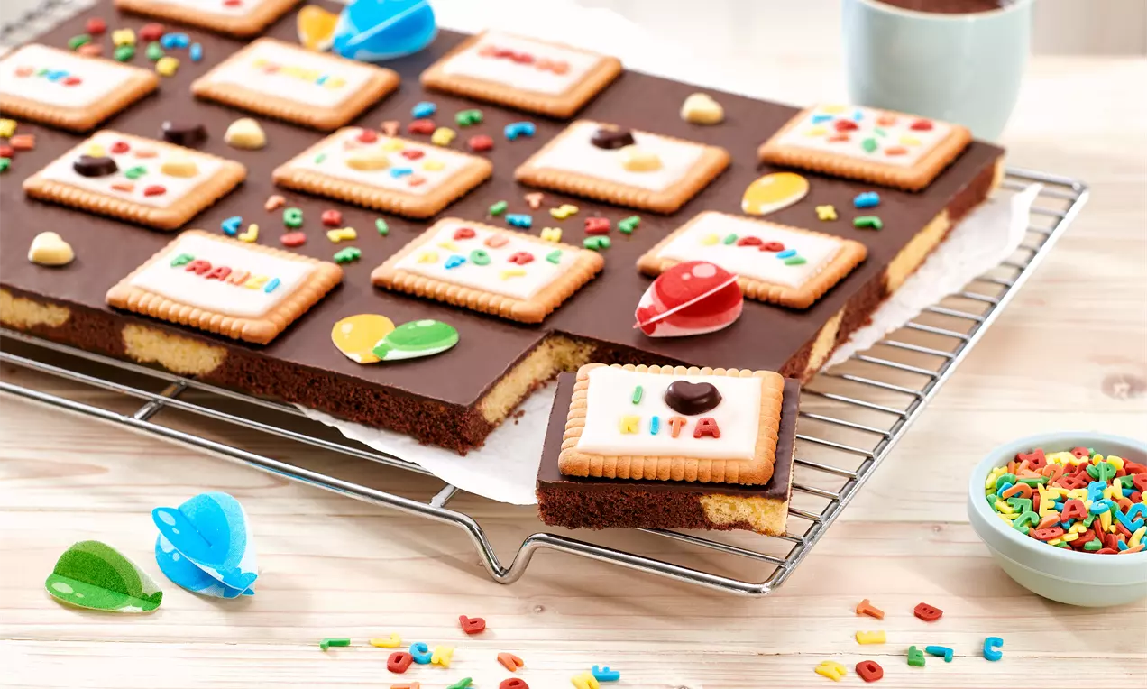 Bunter Blechkuchen mit Schokoglasur, belegt mit Keksen, Zuckerdekor und Farbperlen, auf einem Gitter mit Spielzeug und Streudeko drapiert