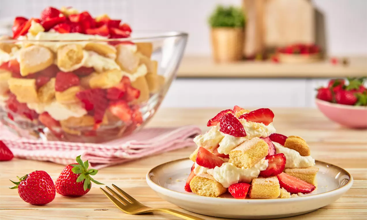 Dessert aus goldgelbem Biskuit, weißer Creme und frischen roten Erdbeeren auf einem Teller, daneben eine Glasschale mit derselben Anordnung