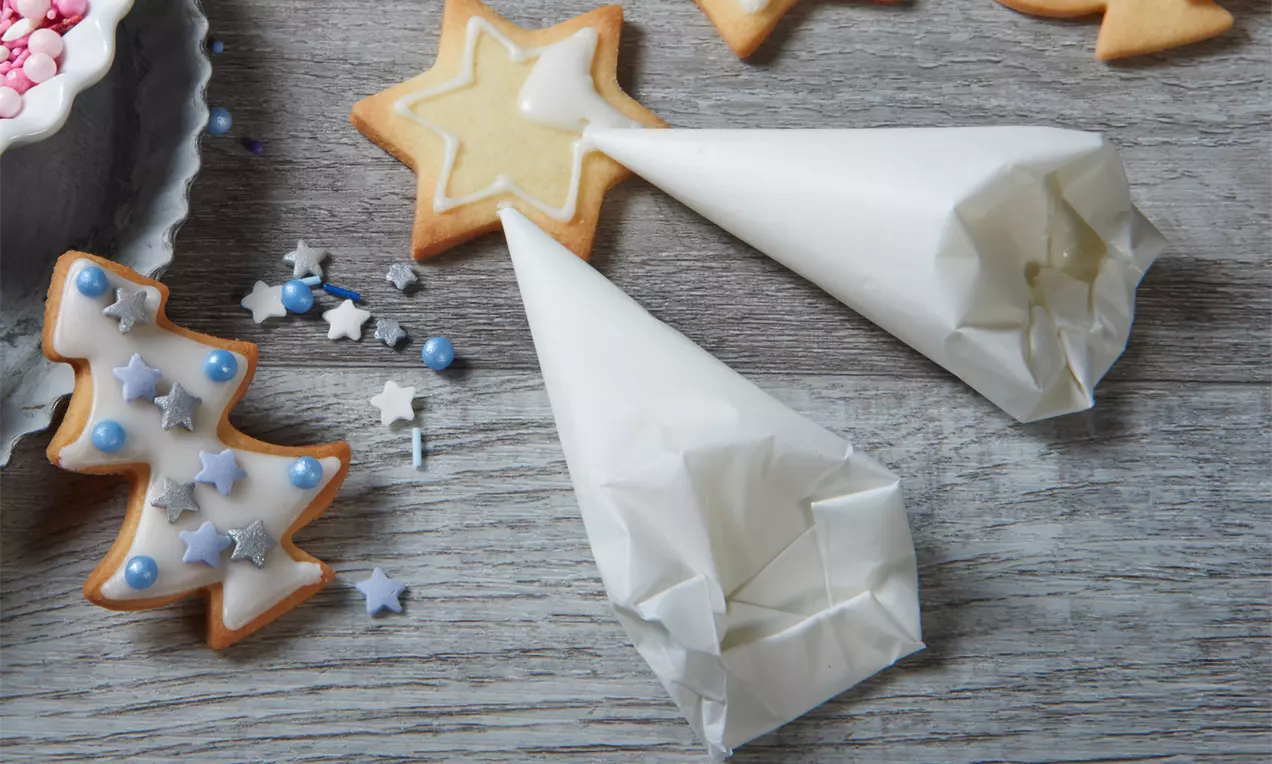 Zwei weiße Papierspritztütchen mit Zuckerguss neben Keksen in Stern- und Baumform auf grauer Holzoberfläche mit Streudekor