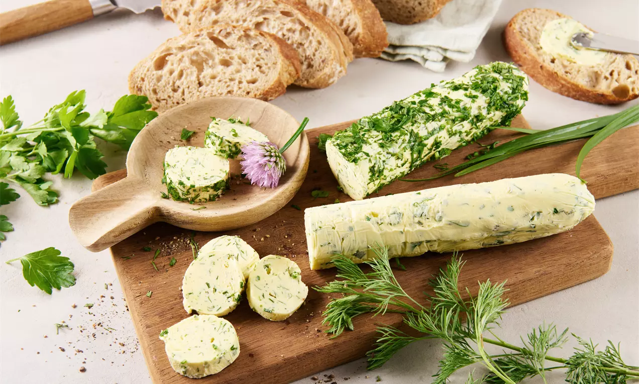 Kräuterbutter in Scheiben geschnitten auf einem hölzernen Brett, garniert mit frischen Kräutern, daneben Brot und Messer drapiert