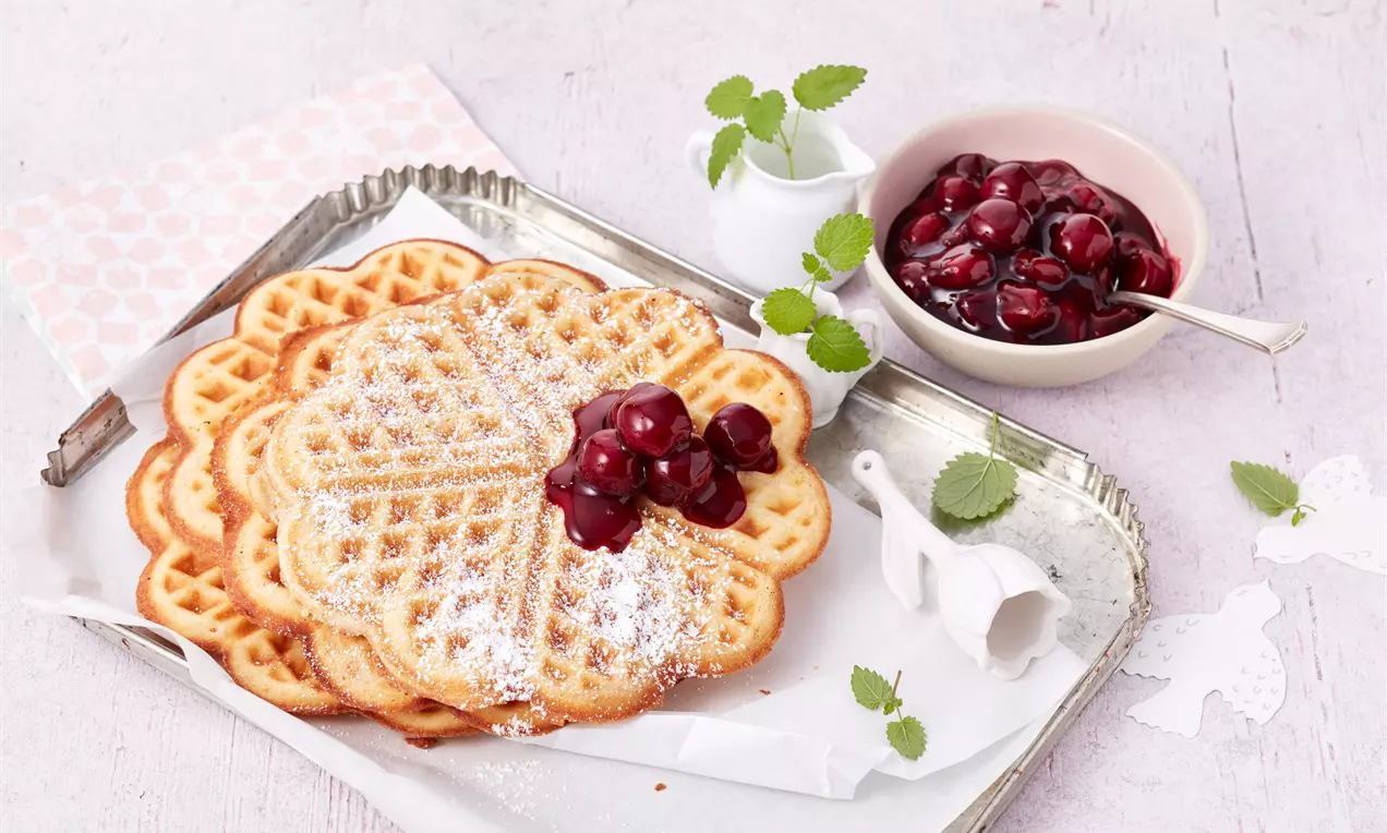 Herzförmige, goldbraune Sahne-Waffeln bestäubt mit Puderzucker, garniert mit glänzenden roten Kirschen auf einem Tablett aus Metall