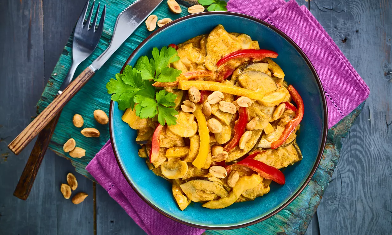 Veganes Gemüsecurry in einer türkisfarbenen Schale mit gelber cremiger Sauce, roten und gelben Gemüsestreifen, Erdnüssen und Kräutern