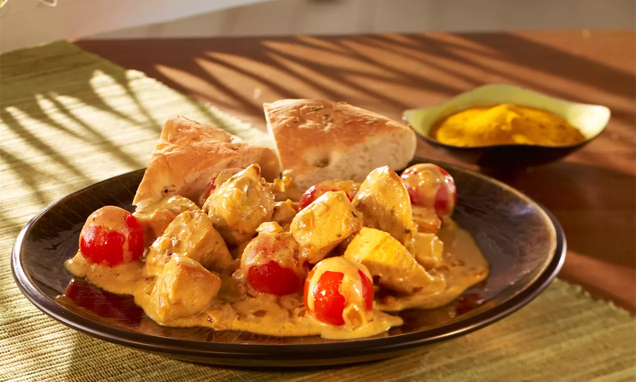Cremiges Hähnchen-Curry mit goldgelber Soße, roten Tomaten und Hähnchenstücken auf dunklem Teller, serviert mit Fladenbrot und Currypulver in Schale