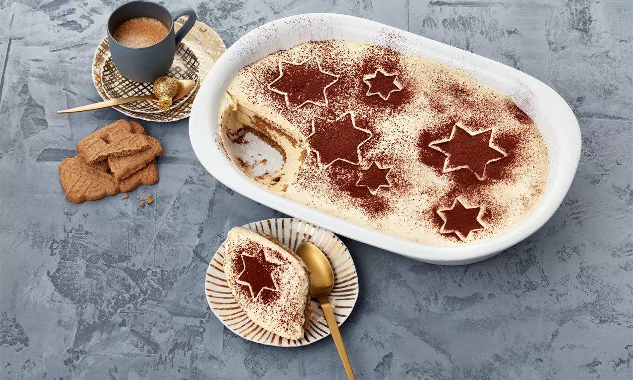 Spekulatius-Tiramisu in weißer Auflaufform mit braunen Kakao-Sternen dekoriert, auf Teller angerichtet neben Keks und Tasse auf grauer Oberfläche