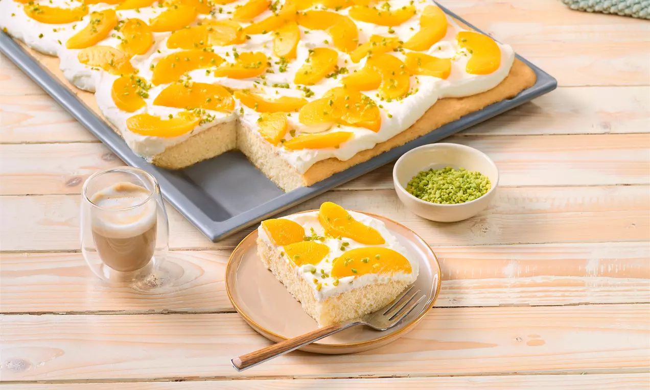 Pfirsichkuchen vom Blech mit Sahnecreme und Pfirsichspalten; ein Stück auf einem Teller serviert, daneben ein Glas Kaffee und eine Schale gehackter Pistazien.