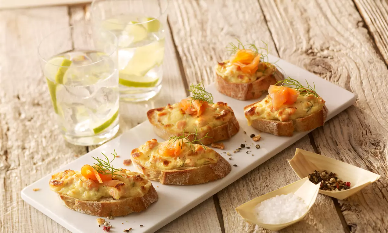 Überbackenes Lachs-Baguette mit rosa Lachs, goldbraunem Käse, cremiger Senf-Joghurt-Honig-Creme, Dill und Zitrone auf rustikalem Holzbrett