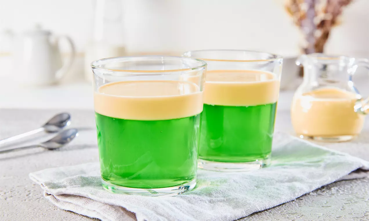Zwei Gläser mit grüner, glänzender Götterspeise und beigefarbener Schicht darauf, auf einem gefalteten Tuch mit kleinen Löffeln daneben