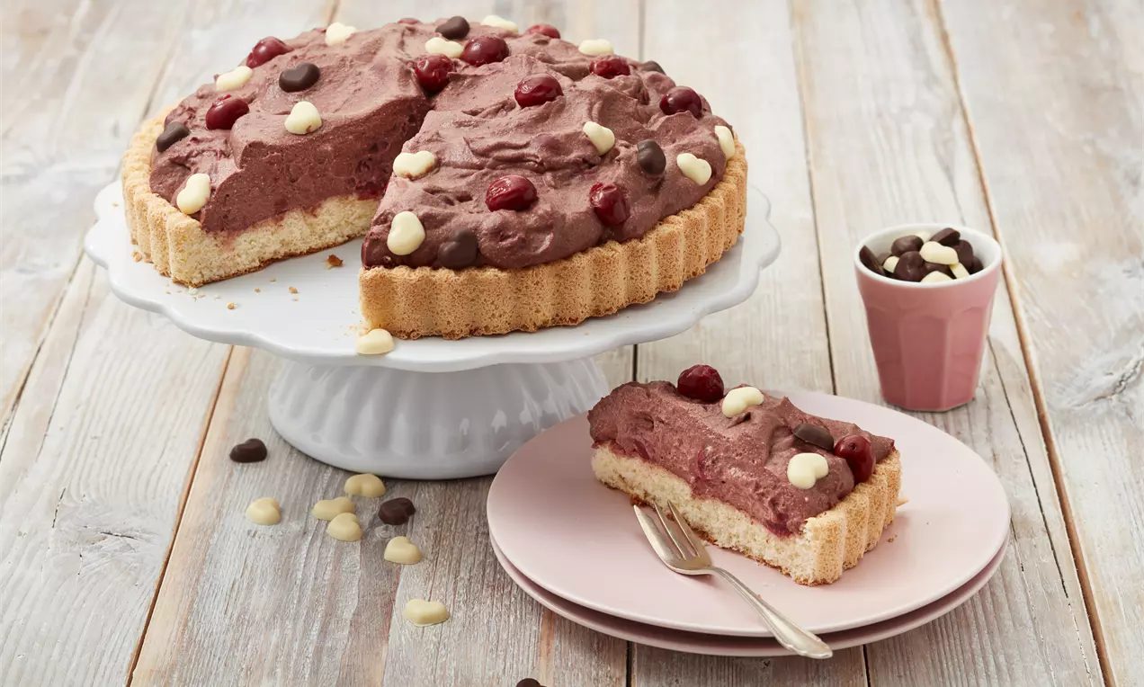 Schokoladiger Schüttelkuchen mit lockerer Creme, Kirschen und zweifarbigen Schokodrops auf Mürbeteigboden, serviert auf weißer Tortenplatte