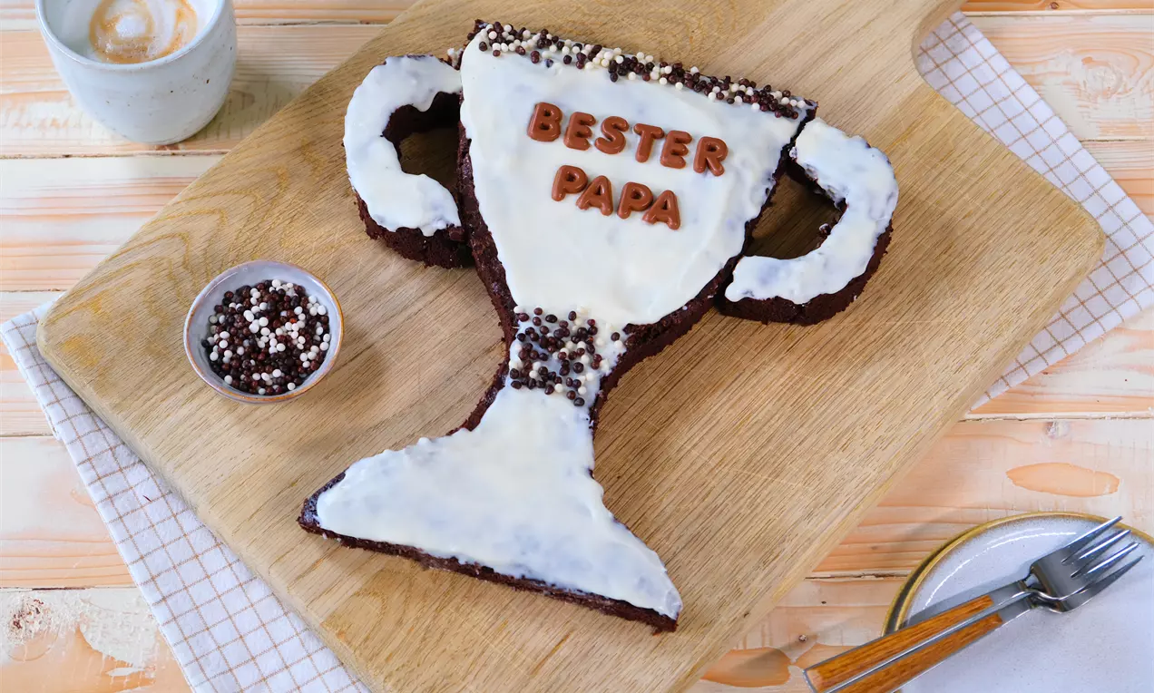 Brauner Brownie in Pokalform mit weißer Glasur, Schokostreuseln und Schriftzug, arrangiert auf Holzbrett mit Besteck und Kaffee in der Umgebung