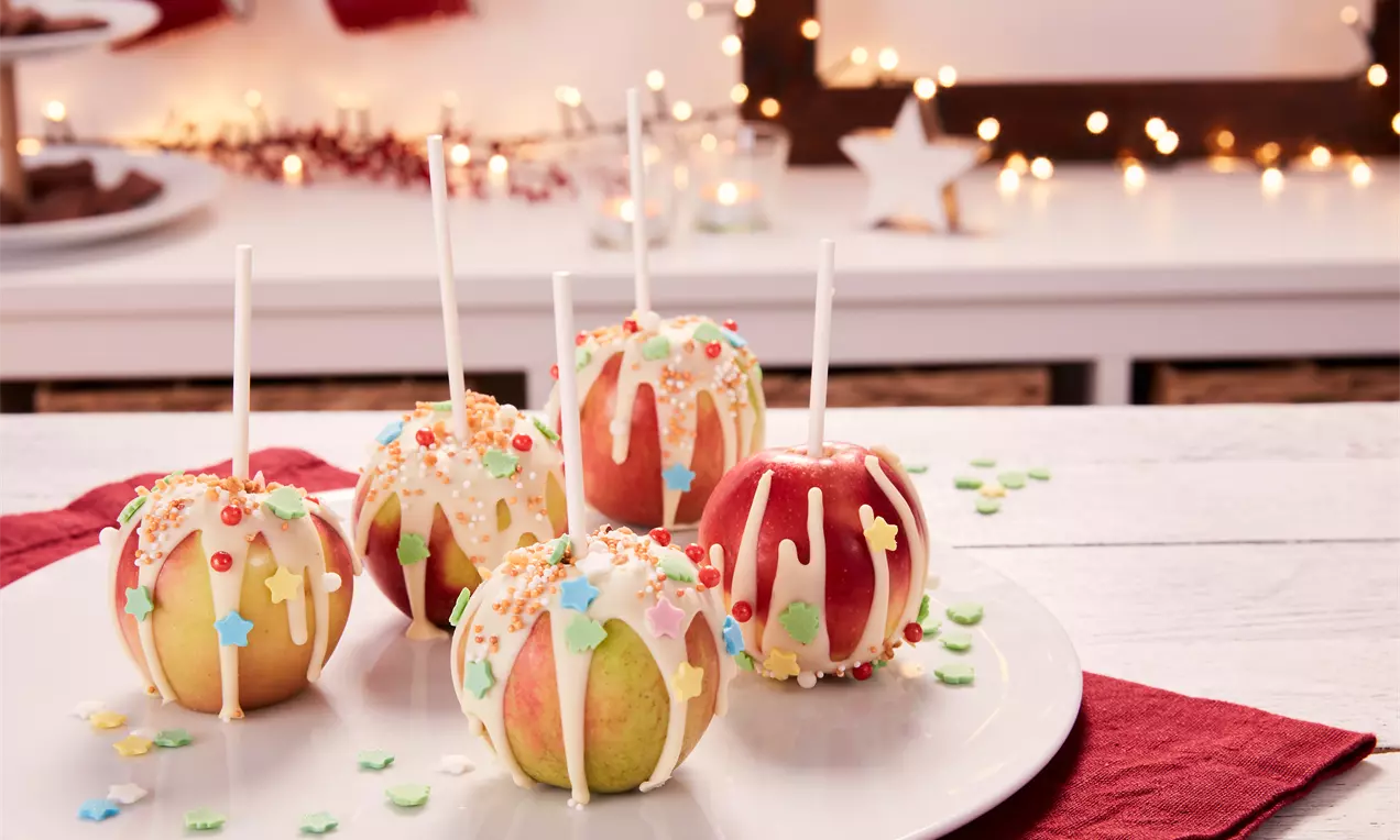 Rote und gelbe Liebesäpfel mit weißem Zuckerguss, bunten Streuseln und Holzstielen auf weißem Teller, umgeben von rotem Stoff und Lichtern