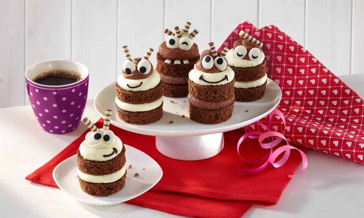 Kleine Schichttörtchen aus dunklem Teig und weißer Creme mit Schokoröllchen-Antennen und Zuckergesichtern auf weißer Etagere drapiert