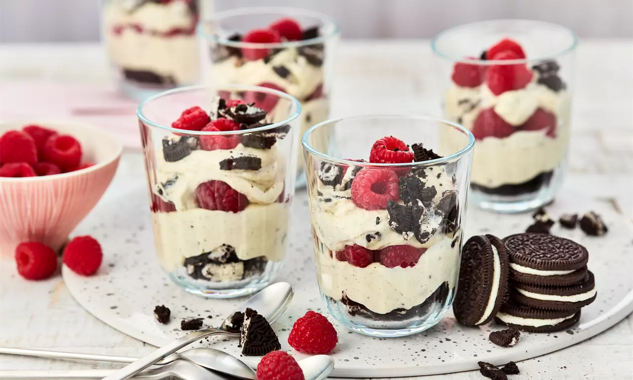 Veganes Oreo-Dessert in fünf Gläsern, geschichtet mit heller Creme, Oreo-Stückchen und frischen Himbeeren. Daneben ganze Oreos, zerbröselte Kekse, Löffel und eine Schale mit Himbeeren.