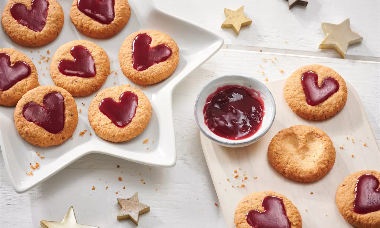 Goldbraune, weiche Plätzchen mit glänzend roter Herzfüllung aus Marmelade auf weißem Sternenteller und Holzbrett mit goldenen Sternen