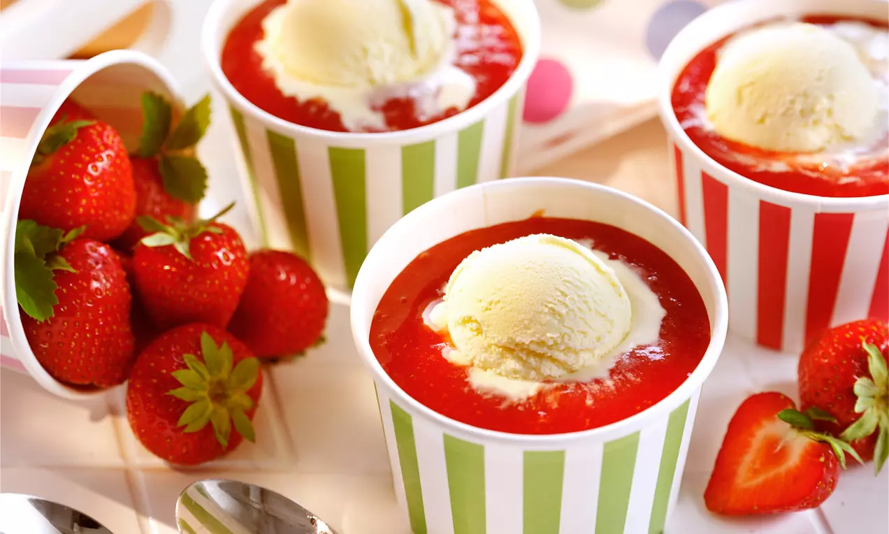 Vanille-Eiskugeln mit glatter Oberfläche in rot-weißer Erdbeersoße, angerichtet in gestreiften Bechern auf Tablett mit frischen Erdbeeren