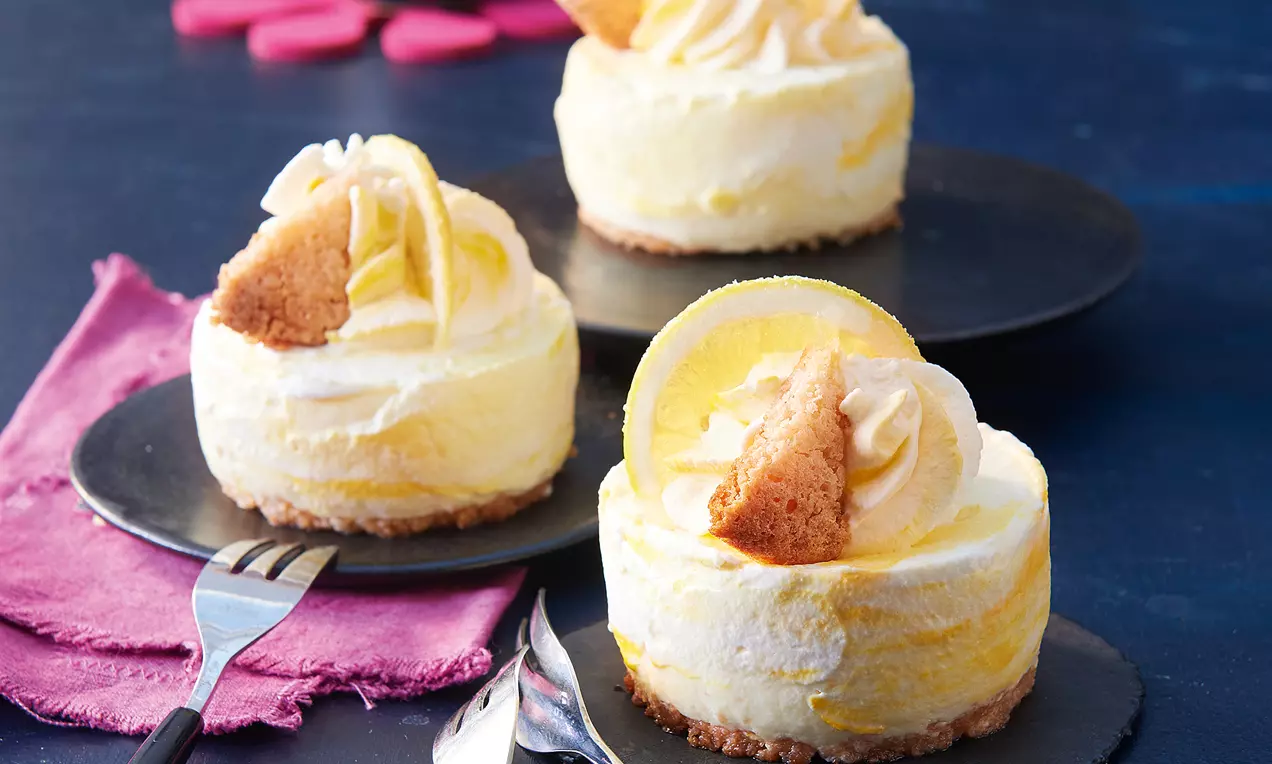 Runde Limoncello-Mousse-Törtchen mit marmorierter Gelb-Weiß-Creme, knusprigem Boden, Sahnehaube und Keksstück auf schwarzen Tellern