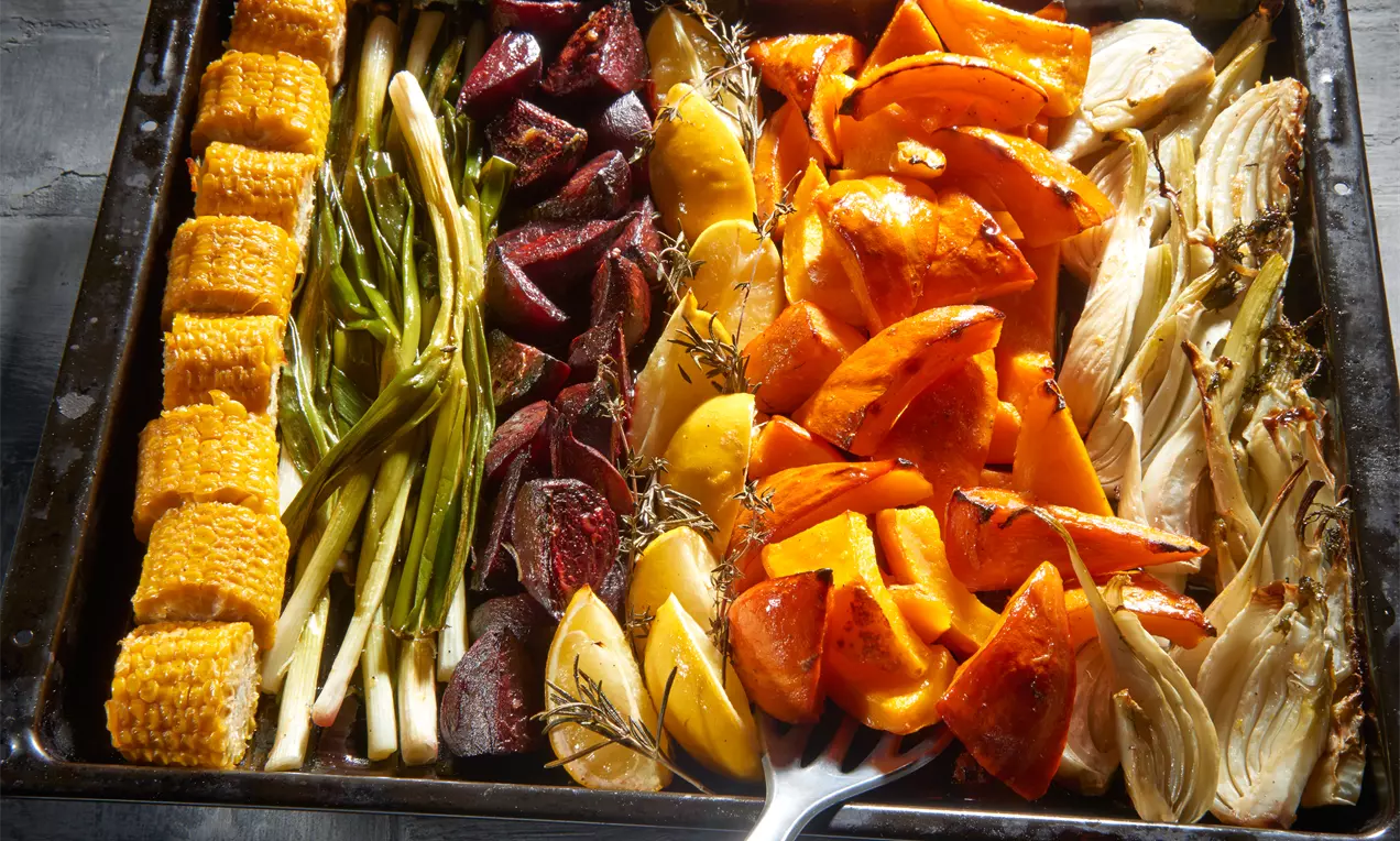 Ofengemüse in Reihen auf einem dunklen Blech, goldgelber Mais, grüner Lauch, rote Rote Bete, gelbe Kartoffeln, orange Kürbis, beigefarbener Fenchel