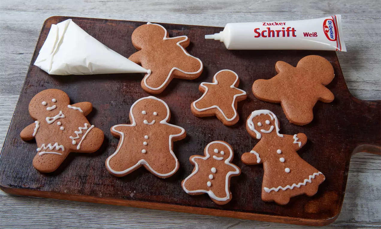 Mehrere braune Lebkuchenfiguren mit weißem Zuckerguss dekoriert auf einem dunklen Holzbrett neben Spritztüte und Zuckerschrift-Tube
