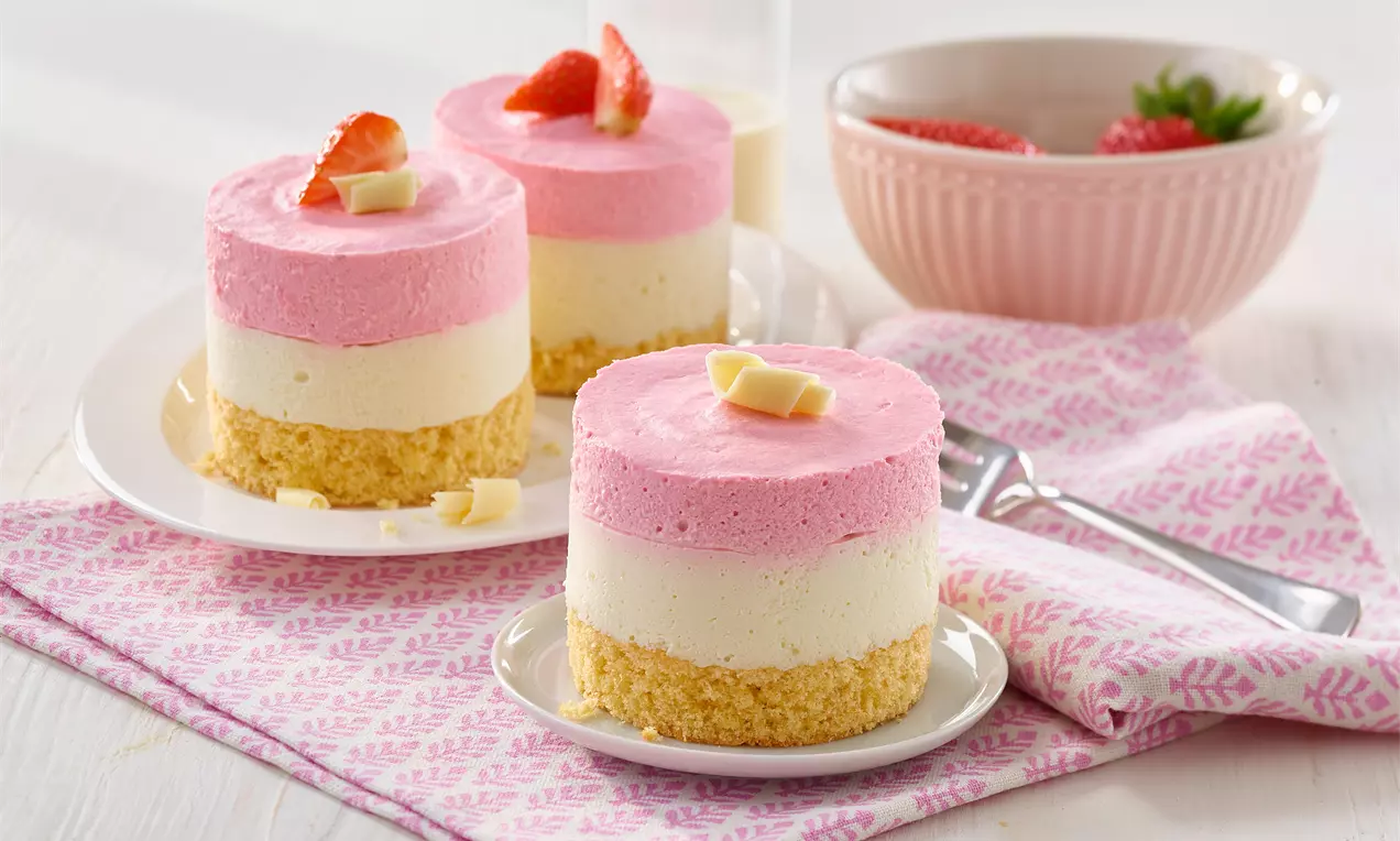 Schicht-Törtchen mit rosa Erdbeercreme, weißer Vanillecreme und hellem Keksboden, garniert mit Erdbeerstück und weißen Schokoladensplittern