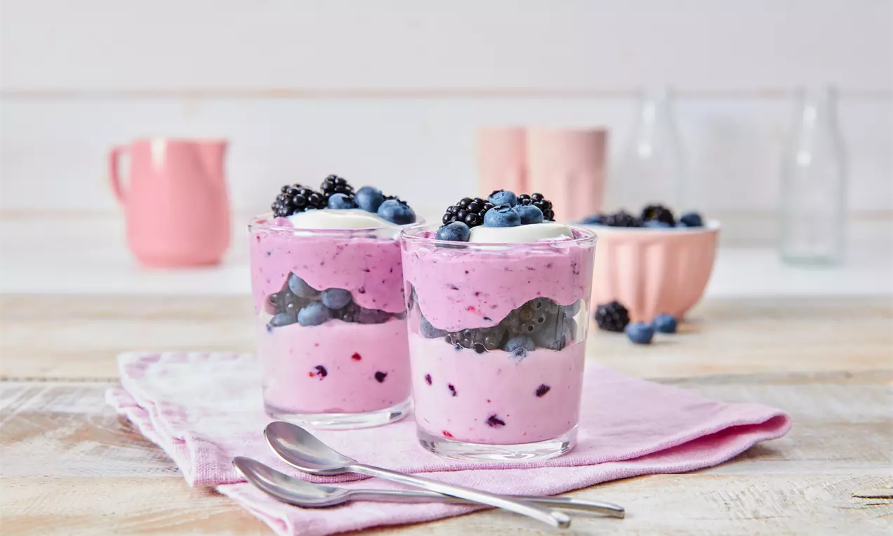 Zwei Gläser mit rosa-cremigem Waldbeeren-Dessert, geschichtet mit Beeren, auf rosa Stofftuch mit zwei Löffeln dekoriert in heller Umgebung
