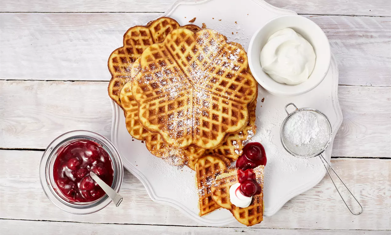 Omas Waffeln: goldbraune, knusprige Waffeln auf weißem Teller, mit Puderzucker bestäubt, frischen roten Erdbeeren und Sahne