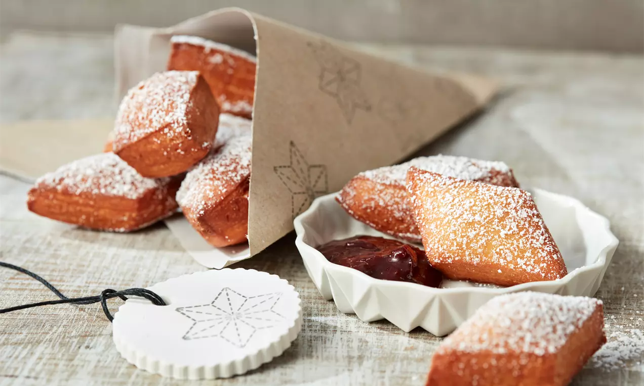 Goldbraune, würfelförmige Schmalzkuchen mit Puderzucker bestäubt, teils in einer weißen Schale mit roter Konfitüre und Papier-Trichter serviert