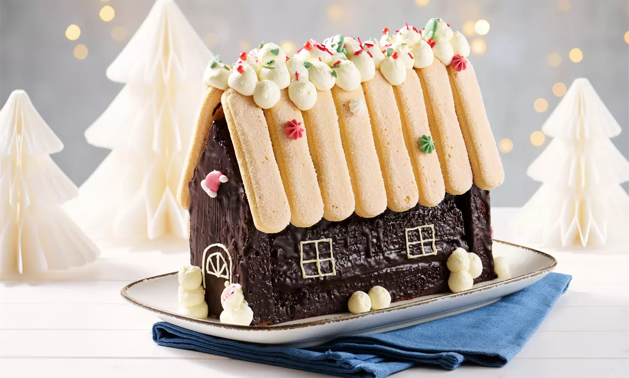 Lebkuchenhaus mit dunkler Schokoladentextur, hellem Biskuitdach, Sahnetupfen und Zuckerstreuseln auf einer Platte vor weißen Papierbäumen