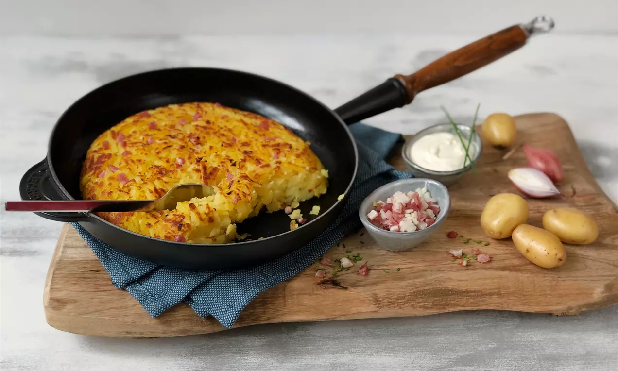 Goldgelber Rösti in einer schwarzen Pfanne auf einem Holzbrett, daneben kleine Kartoffeln, Schmand, Speckwürfel und eine Schalotte drapiert