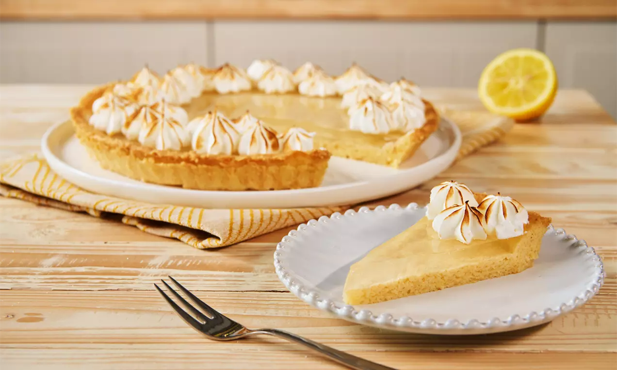Ein Stück goldgelber Tart mit knusprigem Rand und karamellisierten Baiserhauben auf einem weißen Teller neben der ganzen Tart