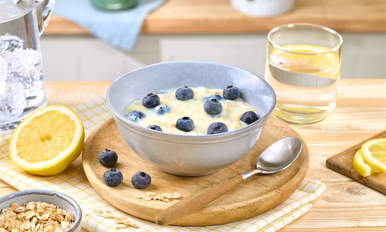 Cremige, vanillegelb schimmernde Pudding-Oats mit frischen, dunkelblauen Heidelbeeren in einer grauen Schale auf hellem Holztablett