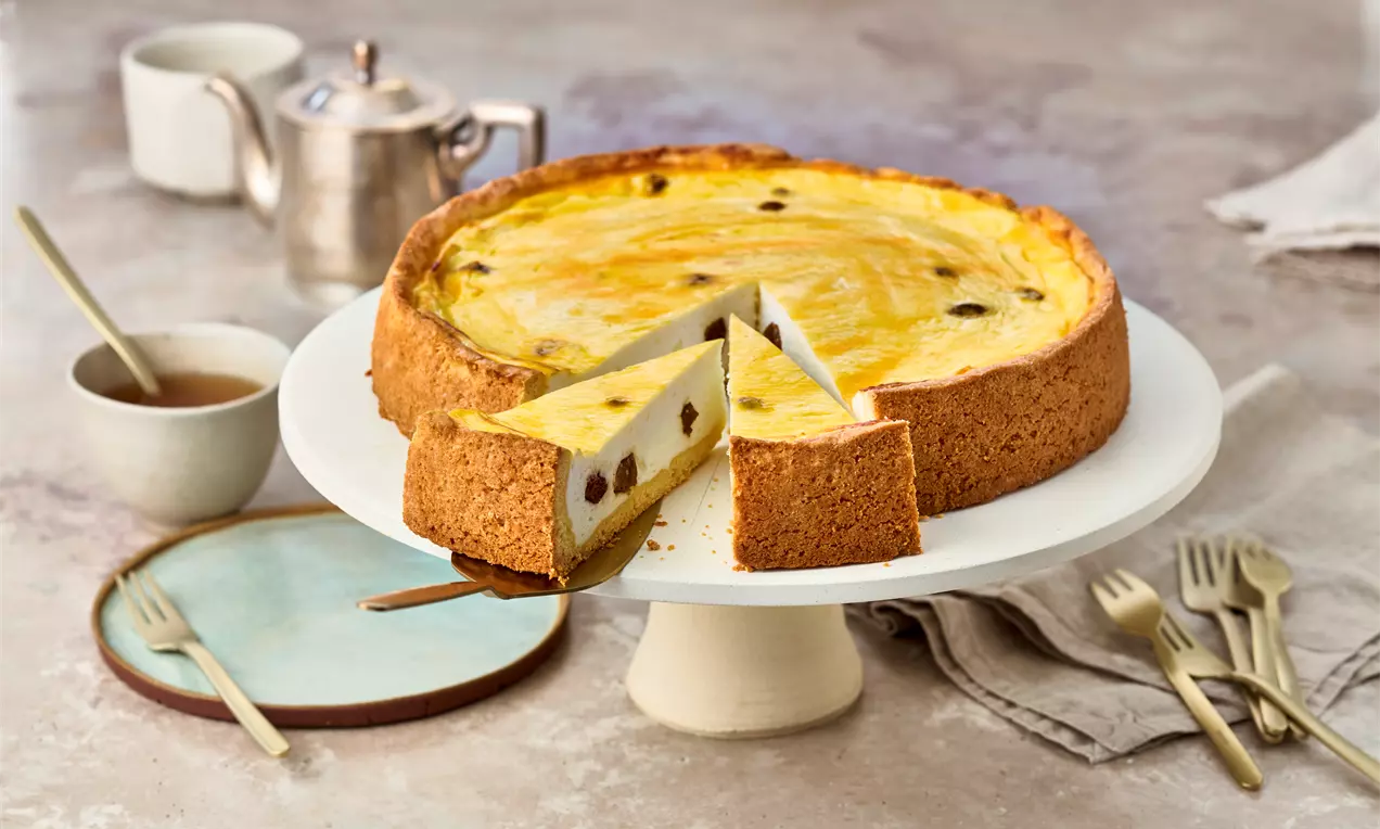 Goldgelber Käsekuchen mit Rosinen in einer hellbraunen, knusprigen Umrandung auf weißer Tortenplatte, angeschnitten mit einer Kuchenheber