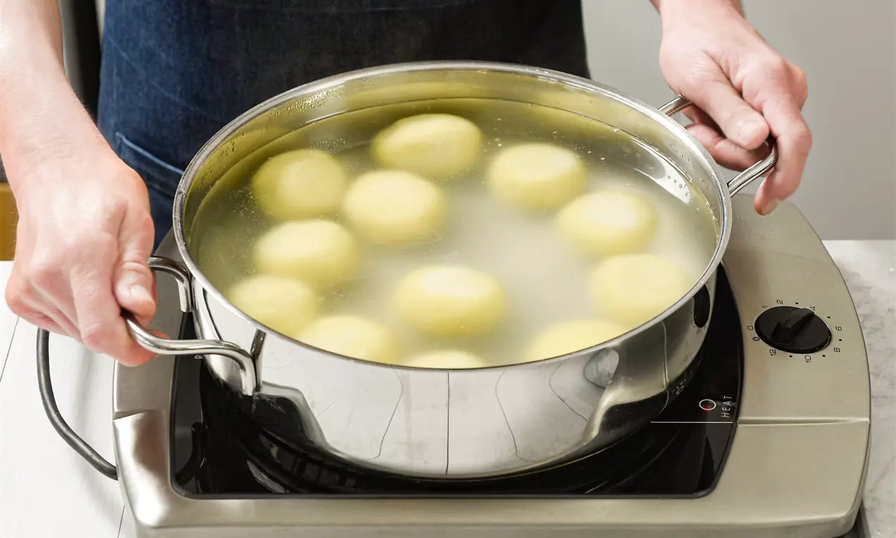 Gelblich-helle Knödel mit glatter Oberfläche garen in leicht trübem Wasser in einem glänzenden Edelstahl-Topf auf einer Kochplatte
