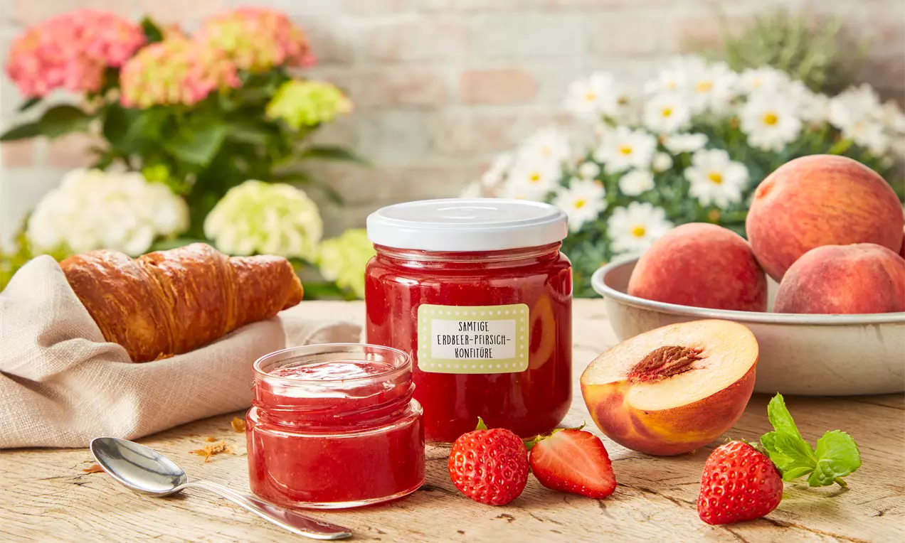 Glas und Schale mit leuchtend roter Erdbeer-Pfirsich-Konfitüre auf einem Holztisch mit frischen Früchten, Croissant und Blütendekor