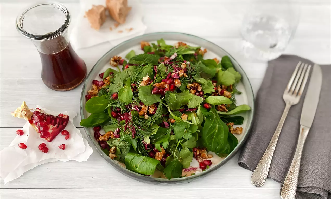 Frischer grüner Salat mit Kräutern, Walnüssen und Granatapfelkernen auf einem runden Glasteller, daneben ein Dressingglas und graues Besteck