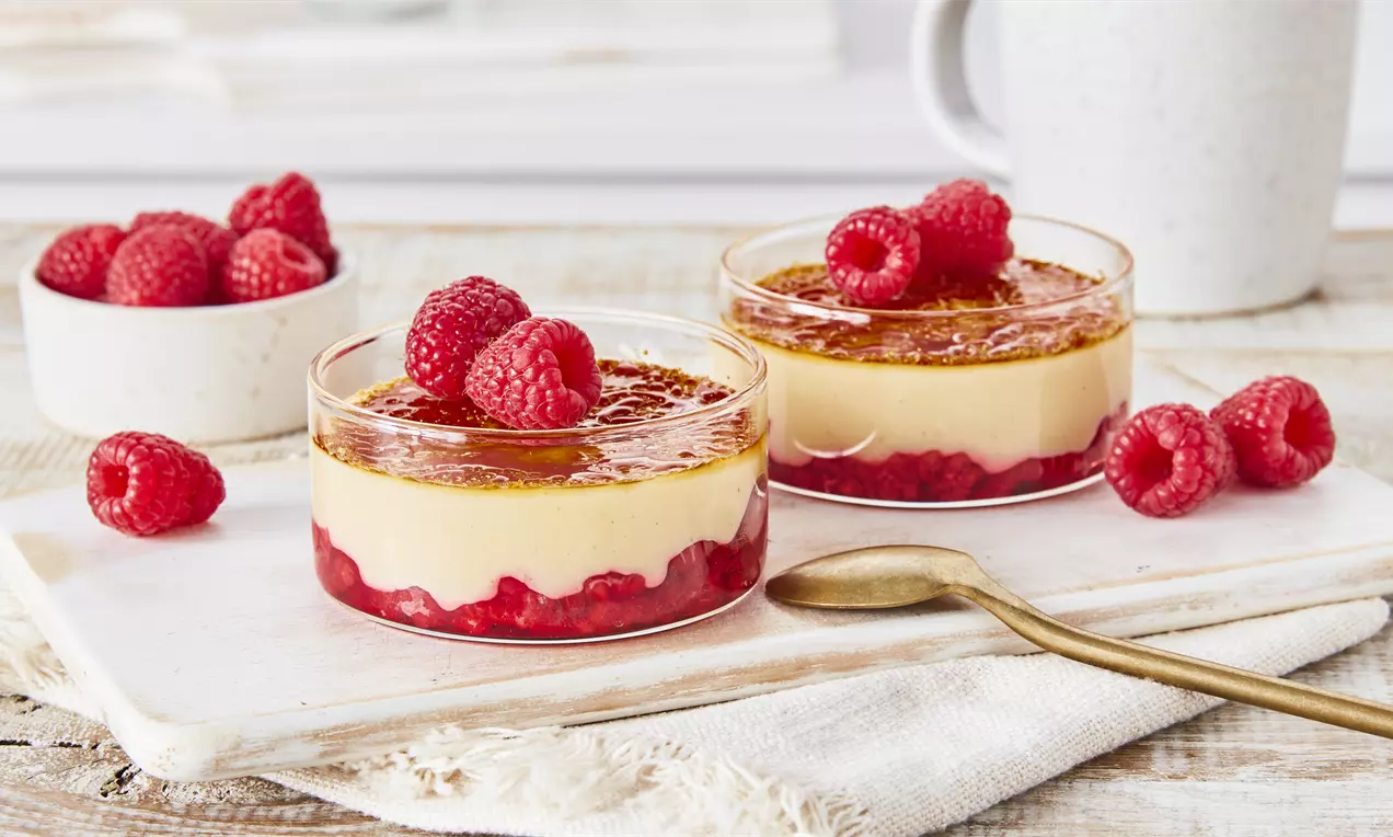 Crème Brûlée mit goldbrauner Karamellkruste und Himbeeren auf roter Fruchtschicht in Glasformen, arrangiert auf hellem Holzbrett