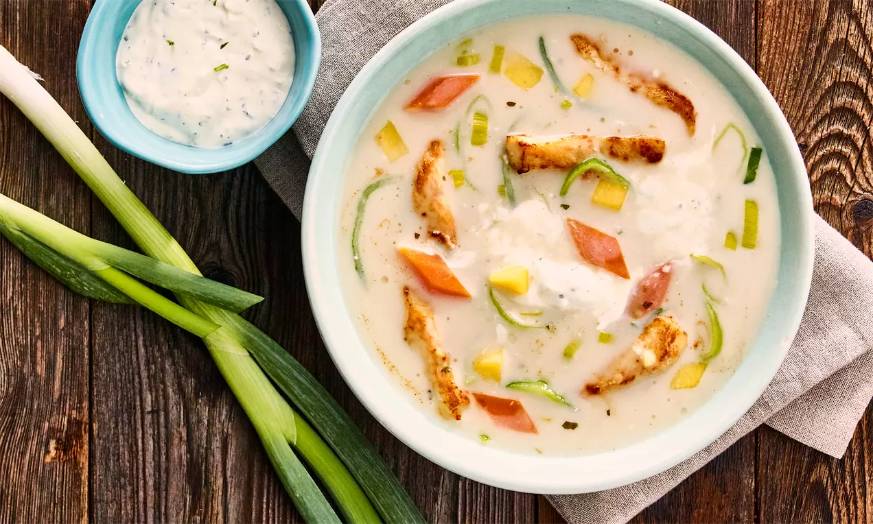 Cremige Geflügel-Kokos-Suppe in einem hellgrünen Schälchen, mit Mango, Lauch, roten Paprikastücken und gebratenen Hähnchenstreifen drapiert
